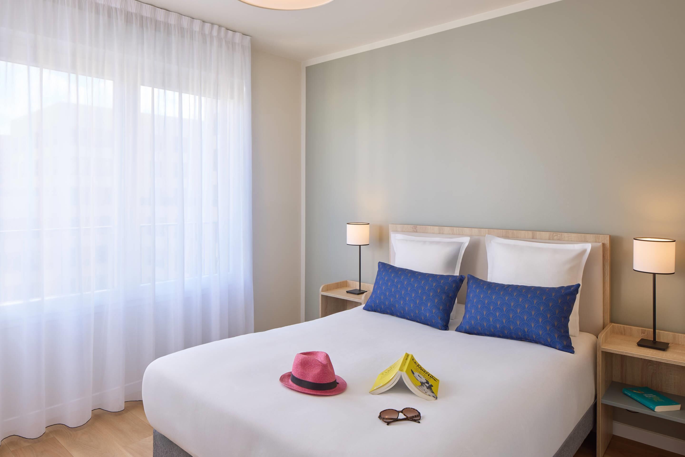 Una cama bien hecha con edredón blanco, dos almohadas, un sombrero rosa, un par de gafas de sol y un libro en la mesita de noche.