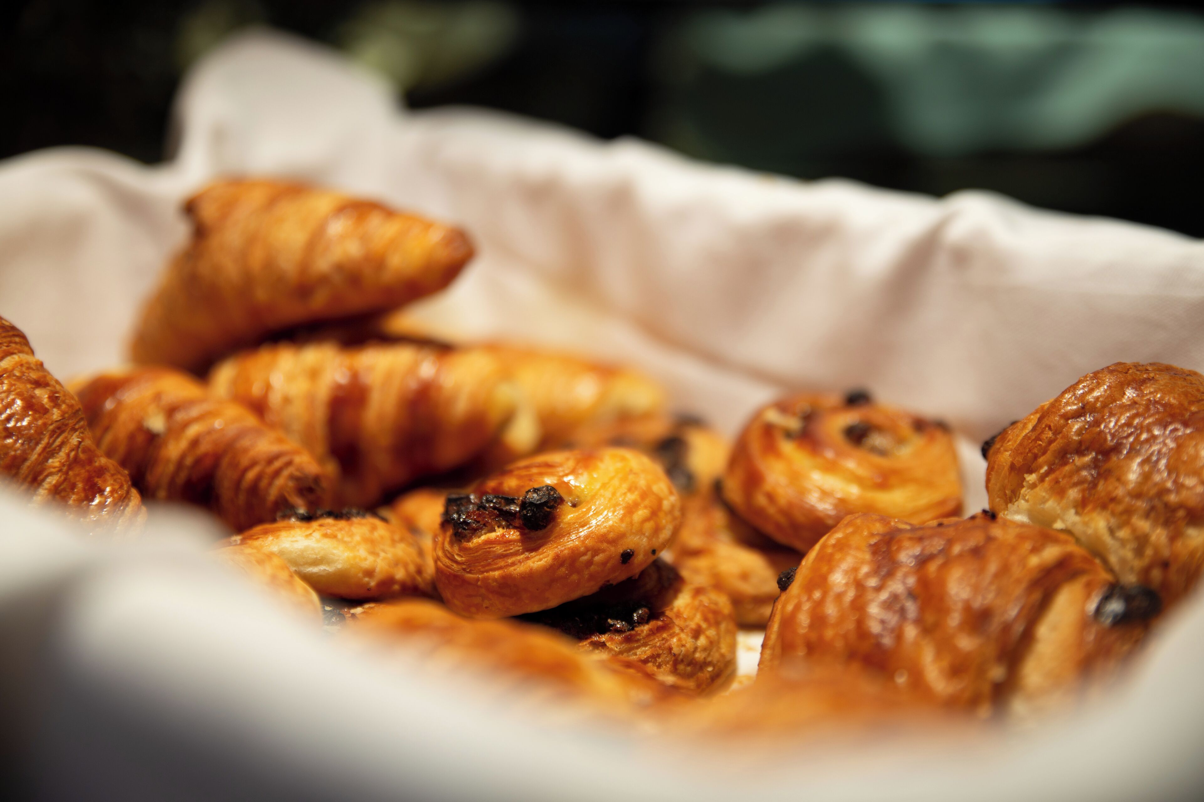Ein Korb mit verschiedenen Backwaren, darunter Croissants und Muffins, auf einer weißen Fläche arrangiert.