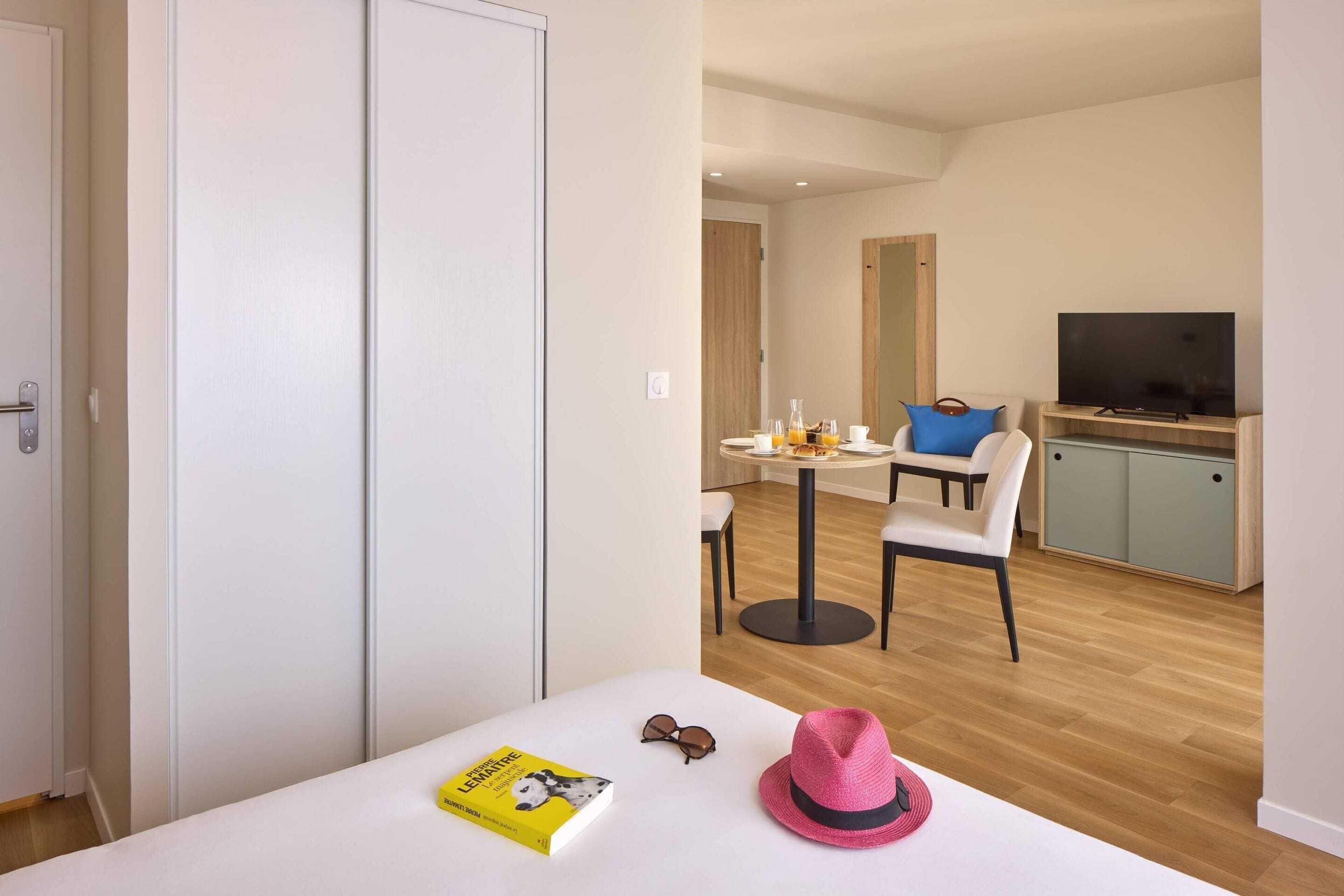 A modern hotel room with a white wardrobe, a dining area with a table and chairs, a television, and a small kitchen area.