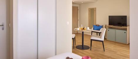 A modern hotel room with a white wardrobe, a dining area with a table and chairs, a television, and a small kitchen area.