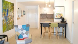 A modern apartment with a living area featuring a colorful armchair, a small dining table with stools, a kitchen with a marble backsplash, and a large abstract painting on the wall. - Esperanza Grand Apartments I • Marbella Center (Marbella)
