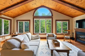 Un salon spacieux avec un haut plafond en bois, de grandes fenêtres, une cheminée et un téléviseur.