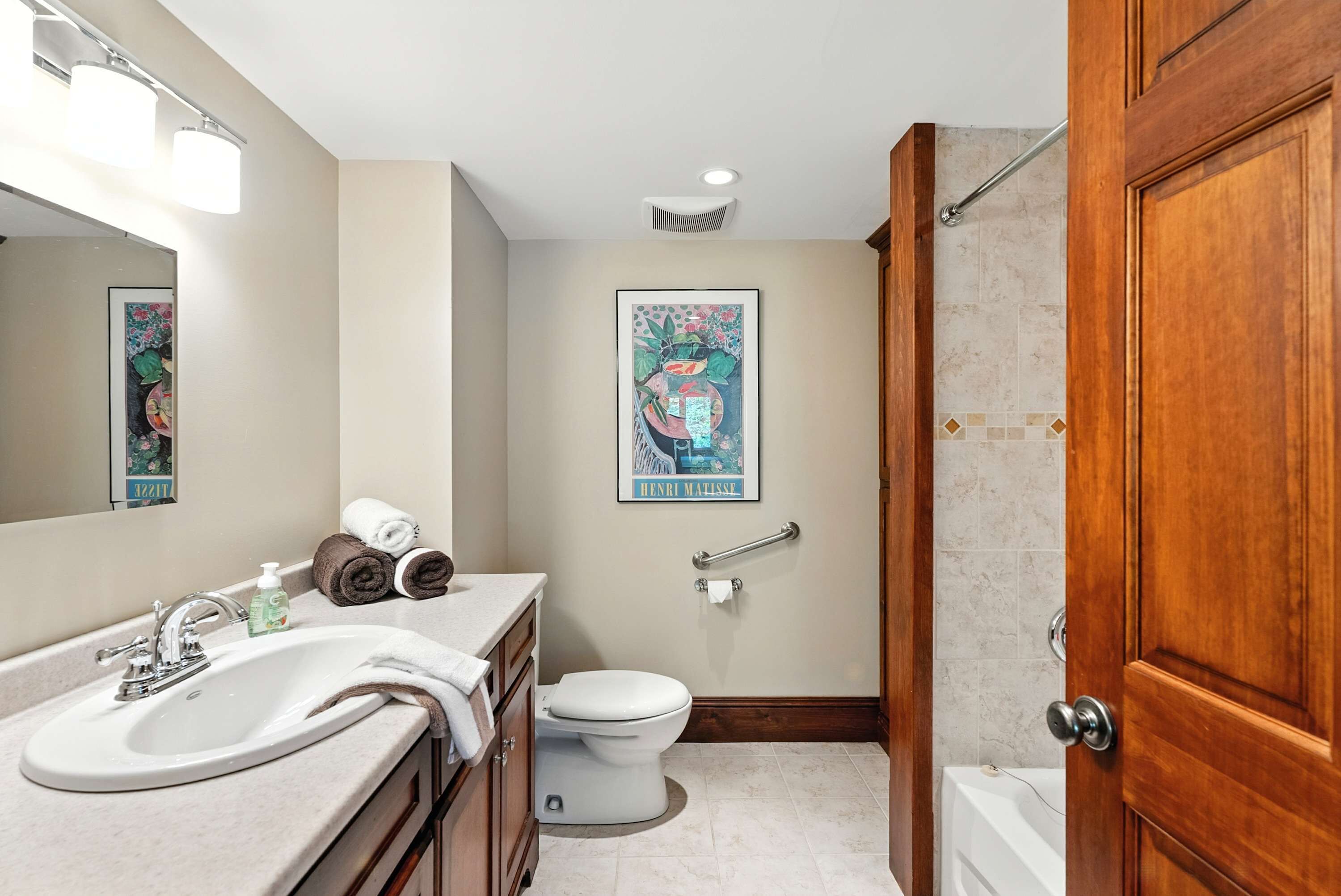 A bathroom with a sink, toilet, and bathtub. There is a towel rack with towels, a framed artwork on the wall, and a wooden door.