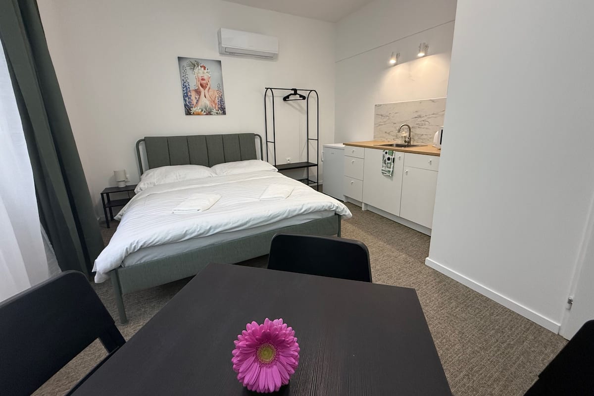 A compact hotel room with a bed, a small table with a flower, a kitchenette, and a window with curtains.