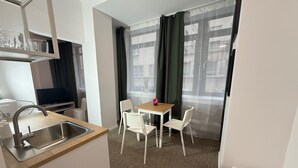A compact hotel room with a kitchenette, a dining area with a table and chairs, and a large window with sheer curtains. - Gremium Apartments (Bratislava)