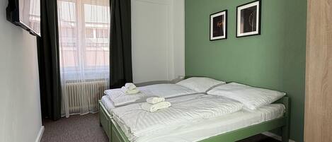 Un dormitorio con pared verde, cama con ropa de cama blanca, un escritorio de madera y un televisor montado en la pared.