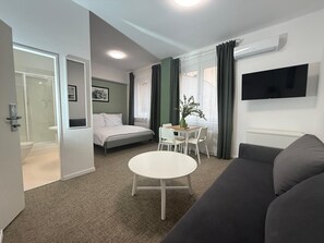 A modern hotel room with a bed, a dining table with chairs, a sofa, a TV mounted on the wall, and a shower area. - Gremium Apartments (Bratislava)
