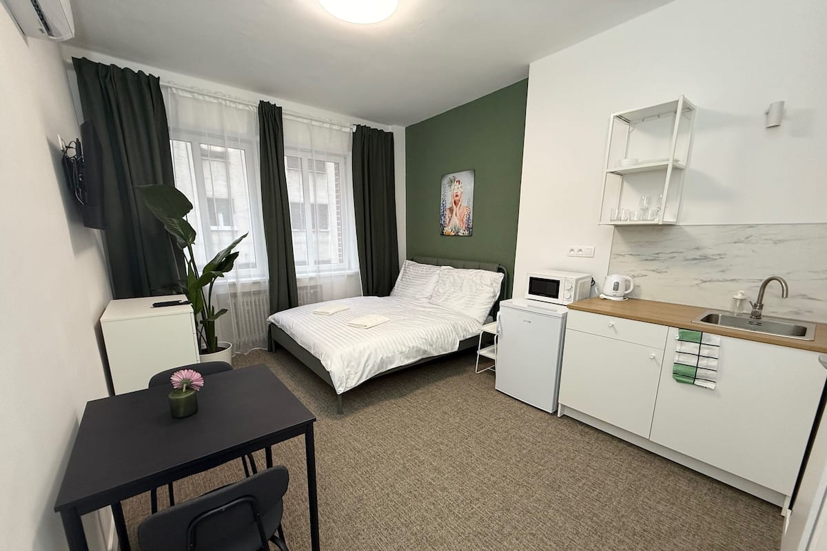 A compact hotel room with a bed, a small kitchenette, a dining area with a table and chair, and a large window with curtains.