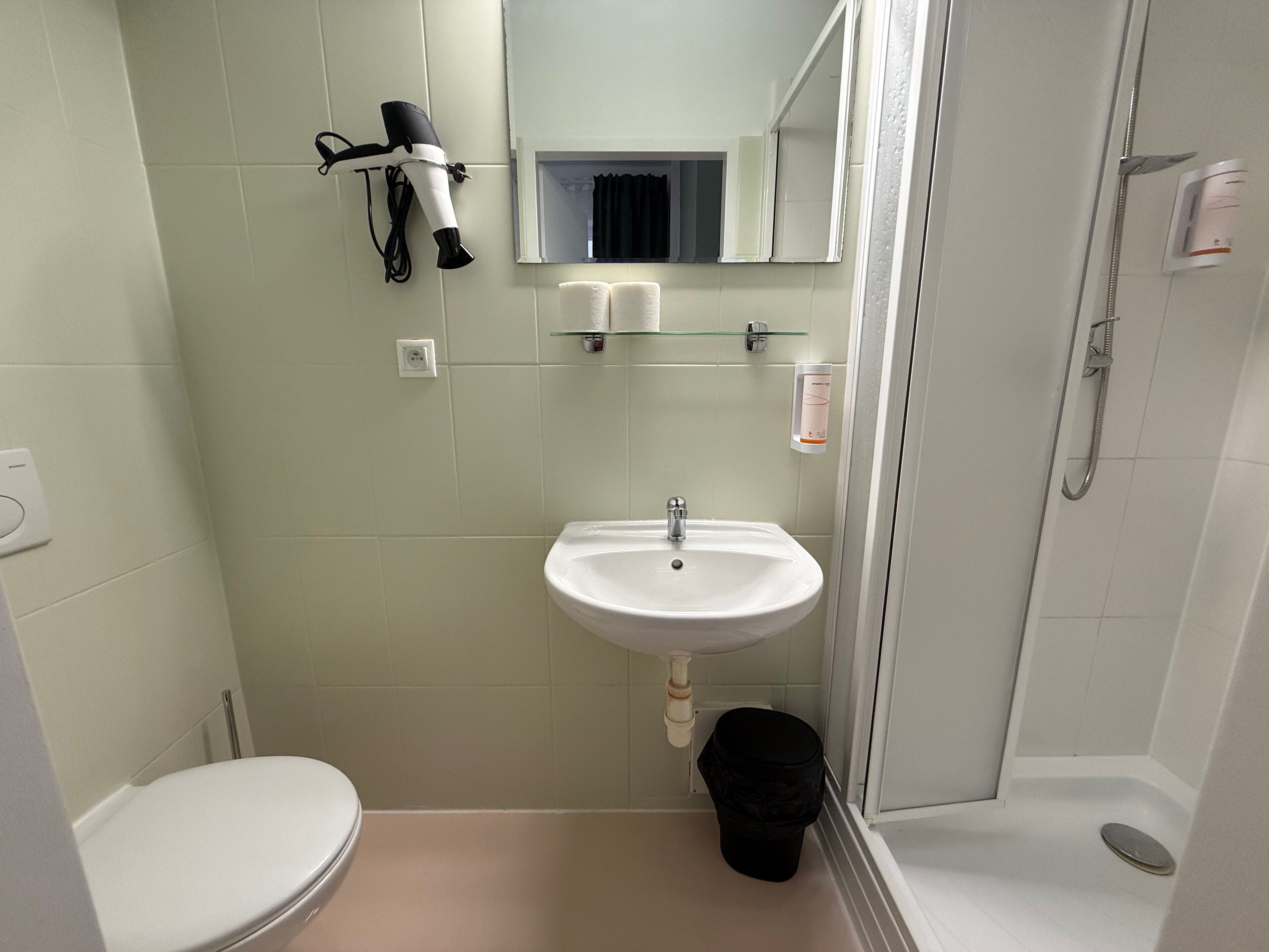 Baño con lavamanos blanco, inodoro, secador de pelo montado en la pared y zona de ducha.