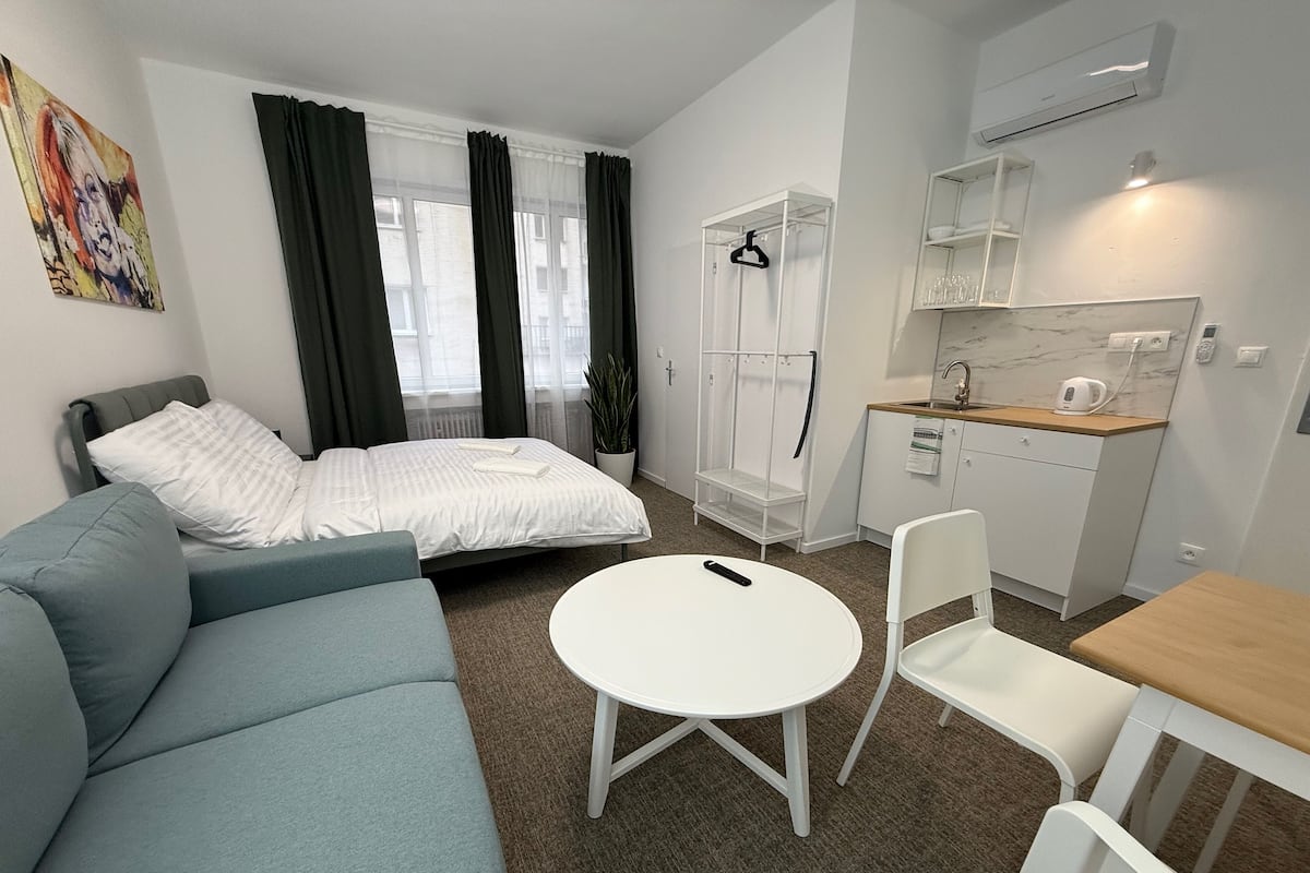 A compact hotel room with a bed, a sofa, a small dining table, and a kitchenette.