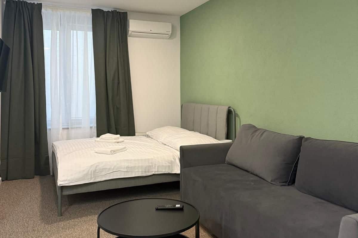 A hotel room with a bed, a sofa, a round black coffee table, and an air conditioning unit on the wall.