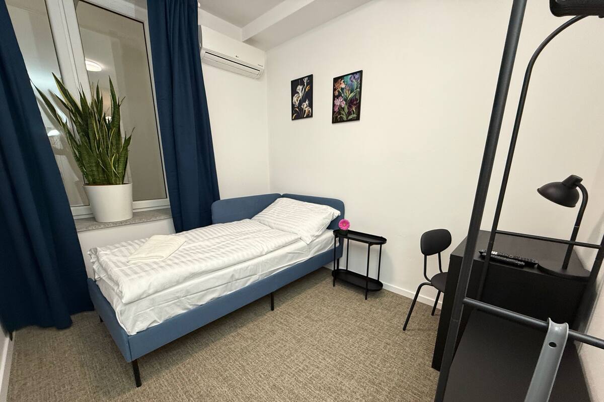 A small, neatly arranged bedroom with a single bed, a desk with a chair, a potted plant, and two wall-mounted paintings.