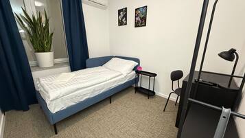 Une petite chambre à coucher, bien rangée, avec un lit simple, un bureau avec une chaise, une plante en pot et deux tableaux accrochés au mur.