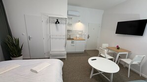 A compact hotel room with a bed, a small kitchenette, a dining area with a table and chairs, a TV mounted on the wall, and a potted plant. - Gremium Apartments (Bratislava)
