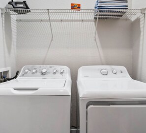 A laundry area with two white front-loading washing machines and a shelf above holding folded towels and a Tide detergent box. - The Epic 00403 Vista Cay Resort Orlando Florida (Orlando)