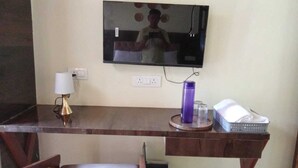 Una habitación con un escritorio de madera, un televisor montado en la pared, una lámpara y un pequeño cubo de basura.