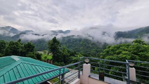 Vista desde un balcón con barandilla de metal, sobre un valle verde y exuberante y montañas cubiertas de niebla.