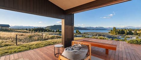 A modern outdoor deck with a wooden bench and a small table, overlooking a scenic landscape with mountains and a body of water.