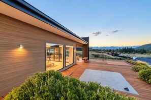 A modern house with a wooden exterior, large glass windows, and a deck with a view of a lake and mountains.