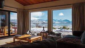 Una sala de estar moderna con techo de madera, ventanas amplias que ofrecen vista a una montaña nevada y un lago, un sofá, una mesa de centro y una cámara sobre trípode.