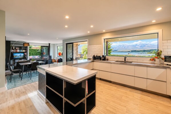 A modern kitchen with a large island, a dining area, and a view of mountains through a large window. - Tasman | 2BR with Fireplace + Hot Tub + BBQ (Lake Tekapo)