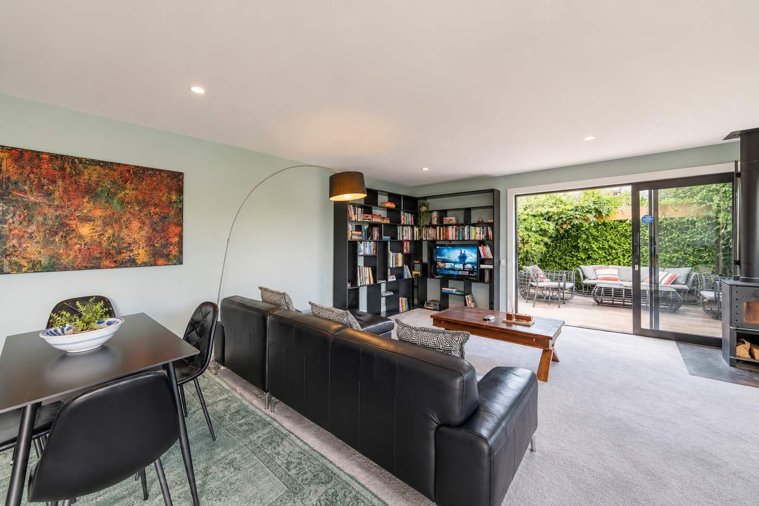 A modern living room with a black leather sofa, a dining table, a bookshelf, and a television.