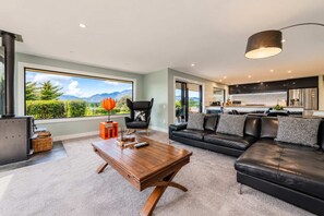 A modern living room with a sofa, a coffee table, and a chair. There is a view of a kitchen and a balcony with a mountain landscape. - Tasman | 2BR with Fireplace + Hot Tub + BBQ (Lake Tekapo)