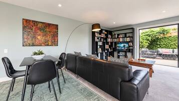 A modern living room with a black leather sofa, a dining table with chairs, a bookshelf, and a large abstract painting on the wall.