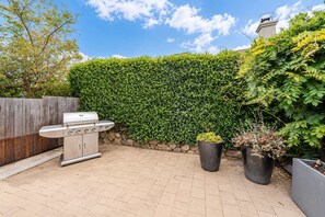 Une terrasse avec un barbecue en acier inoxydable, une clôture en bois, une haie et des plantes en pot.