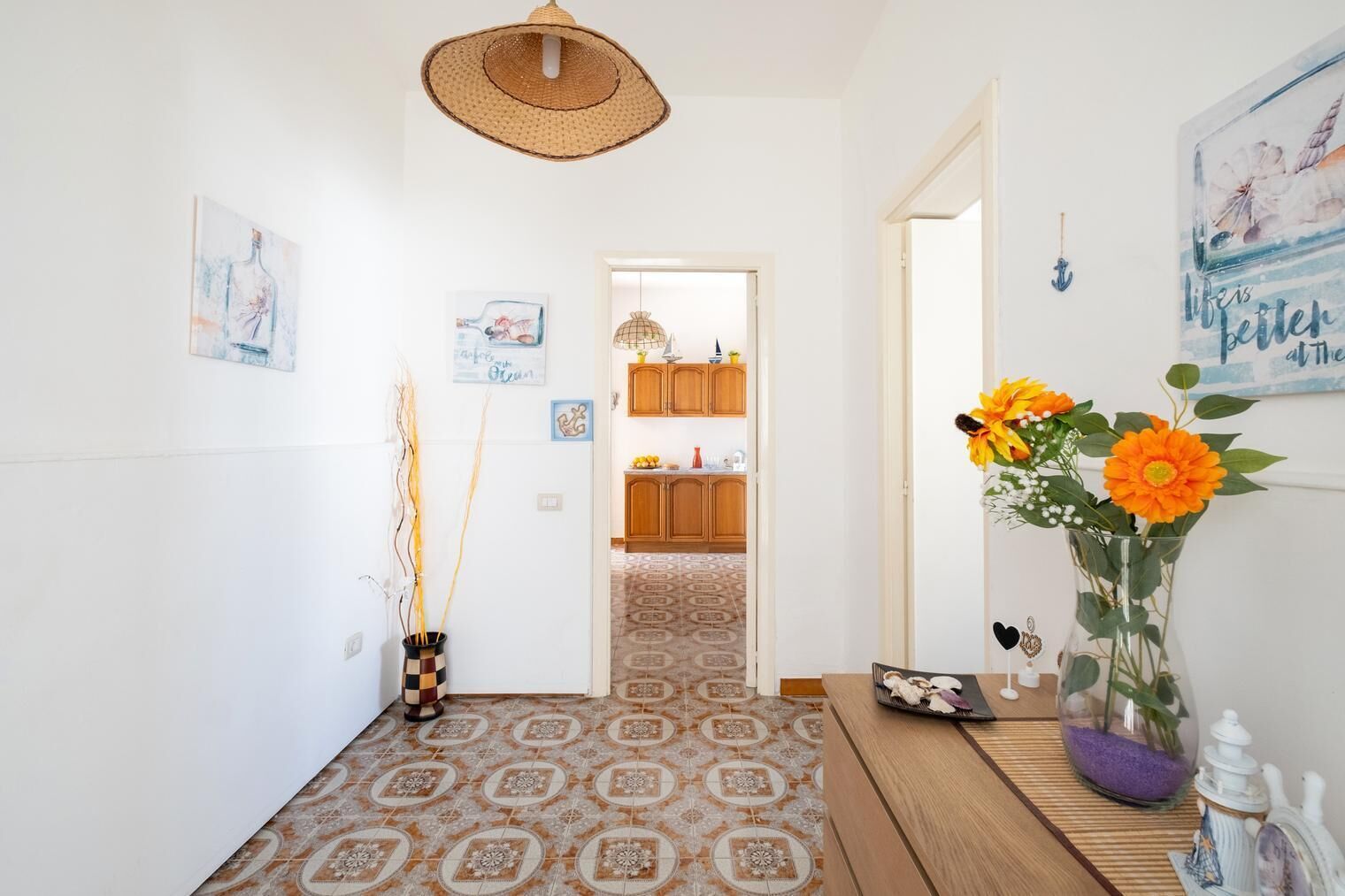 En gang med mønstrede gulvfliser, en vævlampe i loftet og en vase med orange blomster på et træsidebord.