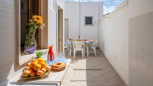 A narrow outdoor patio with a table set for dining, a vase with flowers, and a fruit display.