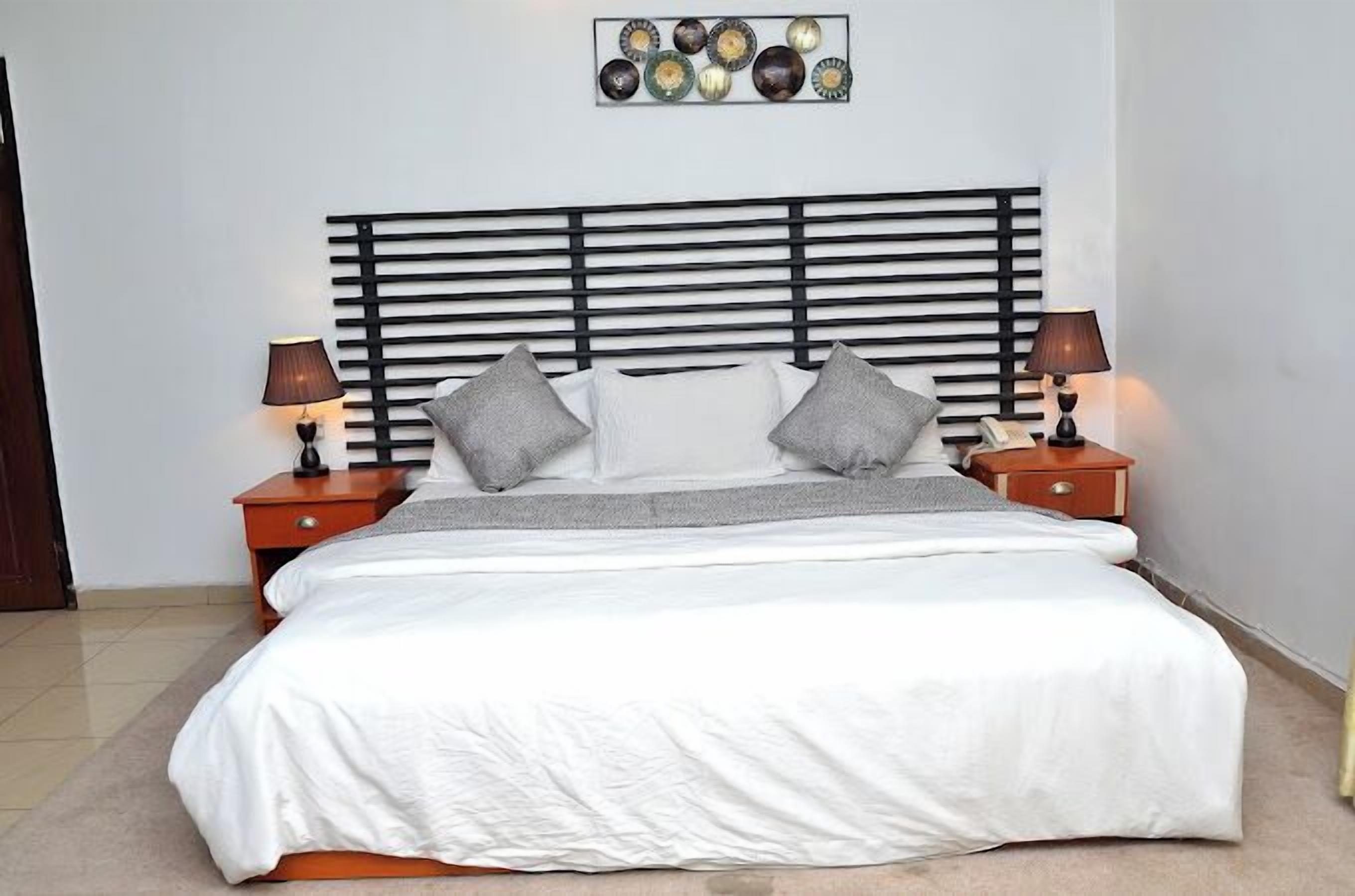 A neatly made bed with a white comforter and gray pillows, flanked by two bedside tables with lamps, set against a white wall with a decorative plate above the bed.
