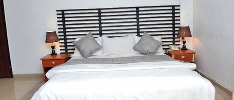 A neatly made bed with a white comforter and gray pillows, flanked by two bedside tables with lamps, set against a white wall with a decorative plate above the bed.