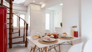 A dining area with a white table set for four, surrounded by white chairs. A wooden spiral staircase is visible to the left.