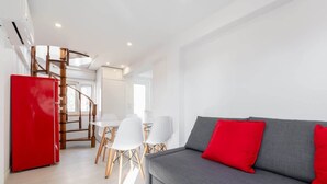 A modern living room with a grey sofa and red cushions, a red refrigerator, and a dining area with white chairs and a wooden table.