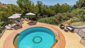 A circular swimming pool with a tiled edge, surrounded by lounge chairs and umbrellas, set against a backdrop of trees and a clear sky.