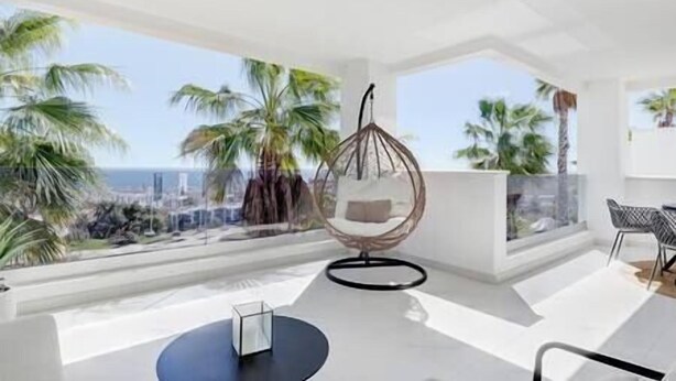 A modern room with a hanging chair, a small table, and a view of palm trees and the sea. - 2 Bedroom Apartment @Estepona Hills With Sea View! (Estepona)