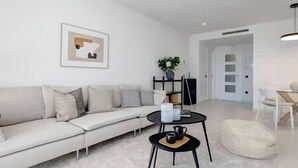 A modern living room with a white sofa, a small black coffee table, and a minimalist decor. - 2 Bedroom Apartment @Estepona Hills With Sea View! (Estepona)