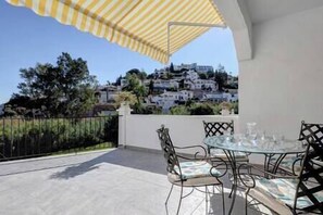 A patio with a glass table and chairs, a yellow striped awning, and a view of white buildings on a hillside.
