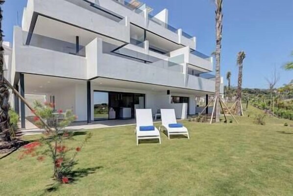 A modern two-story house with a flat roof, large windows, and a covered patio area with two lounge chairs. - Brand New, Luxurious Ground Floor Apartment (Estepona)