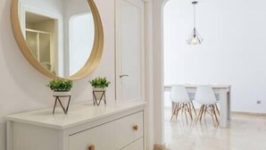 A modern room with a white dresser, a round mirror, two small potted plants, and a dining area with white chairs and a table. - Ground Floor Apartment at Matchroom Country Club (Mijas)