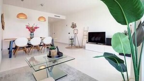 A modern living room with a glass coffee table, a dining area with white chairs, a large green plant, and a flat-screen TV. - Brand New Gorgeous 2 Bed Property @ Estepona Hills (Estepona)