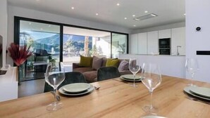 A modern dining area with a wooden table set for four, a sofa, and a view of a mountain landscape through large windows. - Elegant Two Bedroom Apartment at Estepona Hills! (Estepona)