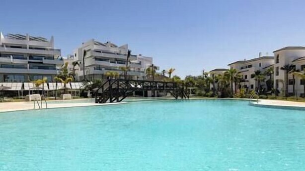 A swimming pool with a bridge structure in the middle, surrounded by modern buildings and palm trees. - Elegant Two Bedroom Apartment at Estepona Hills! (Estepona)
