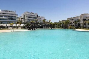 A swimming pool with a bridge structure in the middle, surrounded by modern buildings and palm trees. - Elegant Two Bedroom Apartment at Estepona Hills! (Estepona)