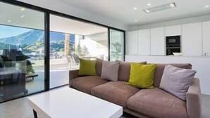 A modern living room with a large window, a sofa, and a kitchen area visible through the glass wall. - Elegant Two Bedroom Apartment at Estepona Hills! (Estepona)