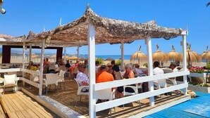A covered outdoor seating area with tables and chairs, overlooking a pool and beach.