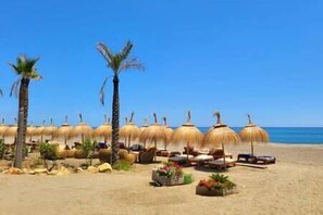 A beach with thatched umbrellas, lounge chairs, and palm trees.