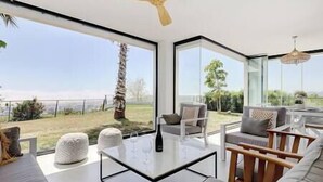 Ein modernes Wohnzimmer mit einem Glastisch, einer Couch und einem Sessel. Ein großes Fenster bietet Blick ins Freie.