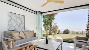 Ein modernes Wohnzimmer mit einer Couch, einem Couchtisch und Sesseln. Ein Deckenventilator, ein großes Fenster mit Blick auf Grünflächen und einen Zaun sind vorhanden.